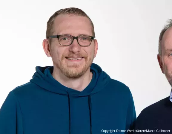 Zwei Männer mittleren Alters und in Pullover schauen lächelnd in die Kamera